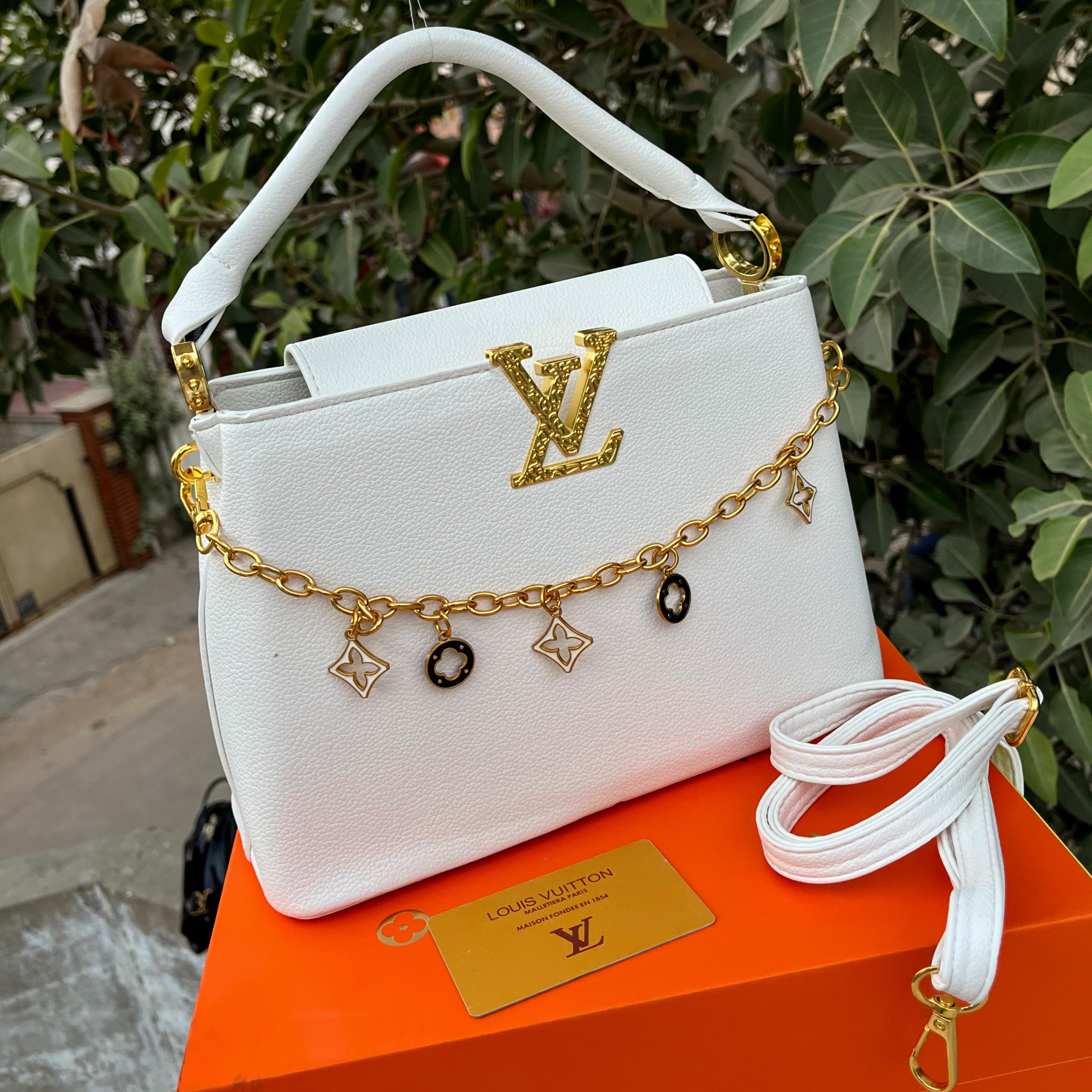 White handbag with gold chain and charms on an orange surface, with a blurred green outdoor background.