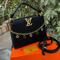 Black handbag with gold chain and charms on an orange Louis Vuitton box against a green leafy background