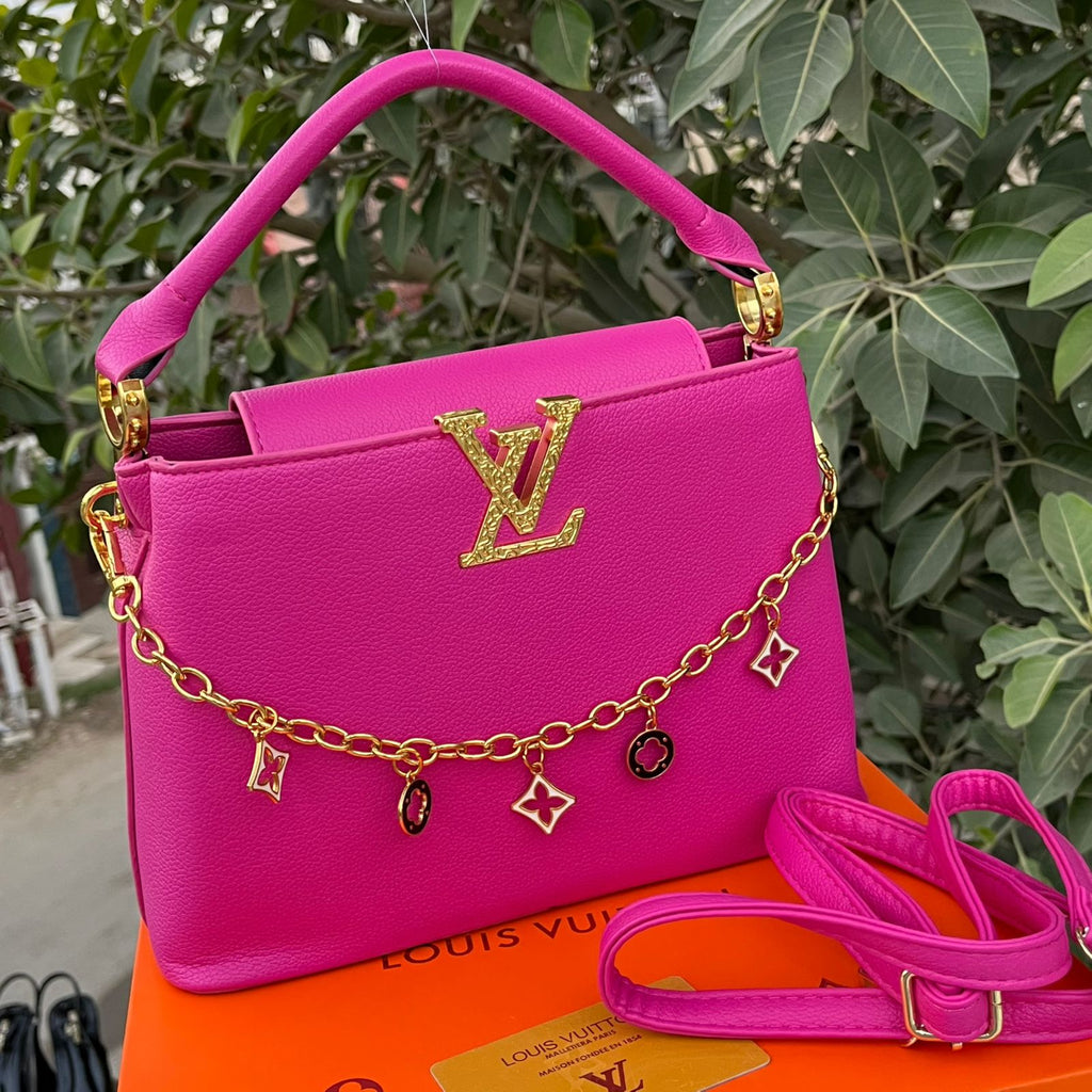 Pink handbag with gold chain and charms on an orange surface with green foliage in the background